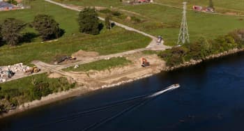 Aerial view of excavators and trucks working along a riverbank while a small boat moves across the water.