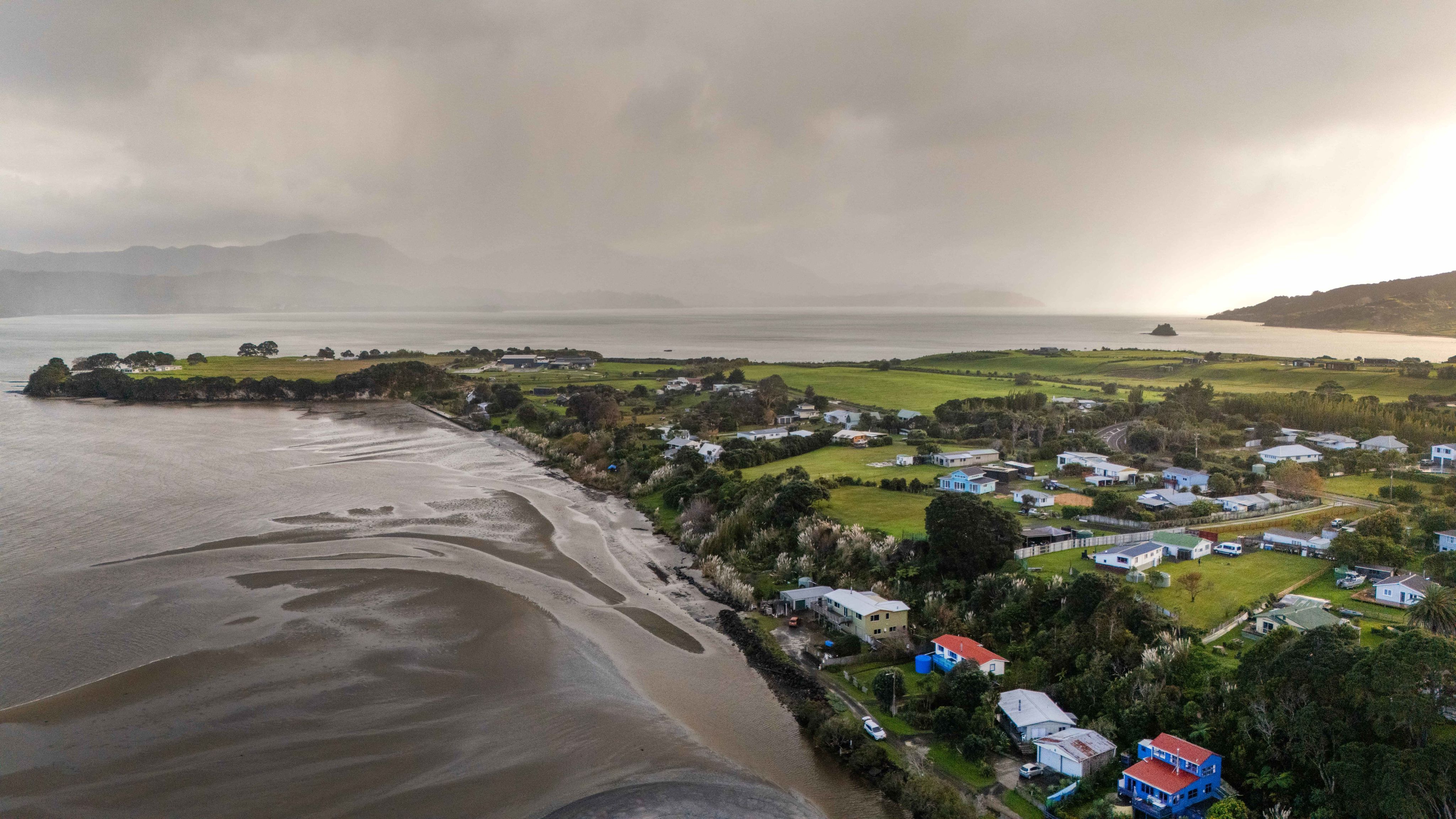 Aerial view of cluster of houses on small peninsula in wide harbour, grey rain clouds behind.