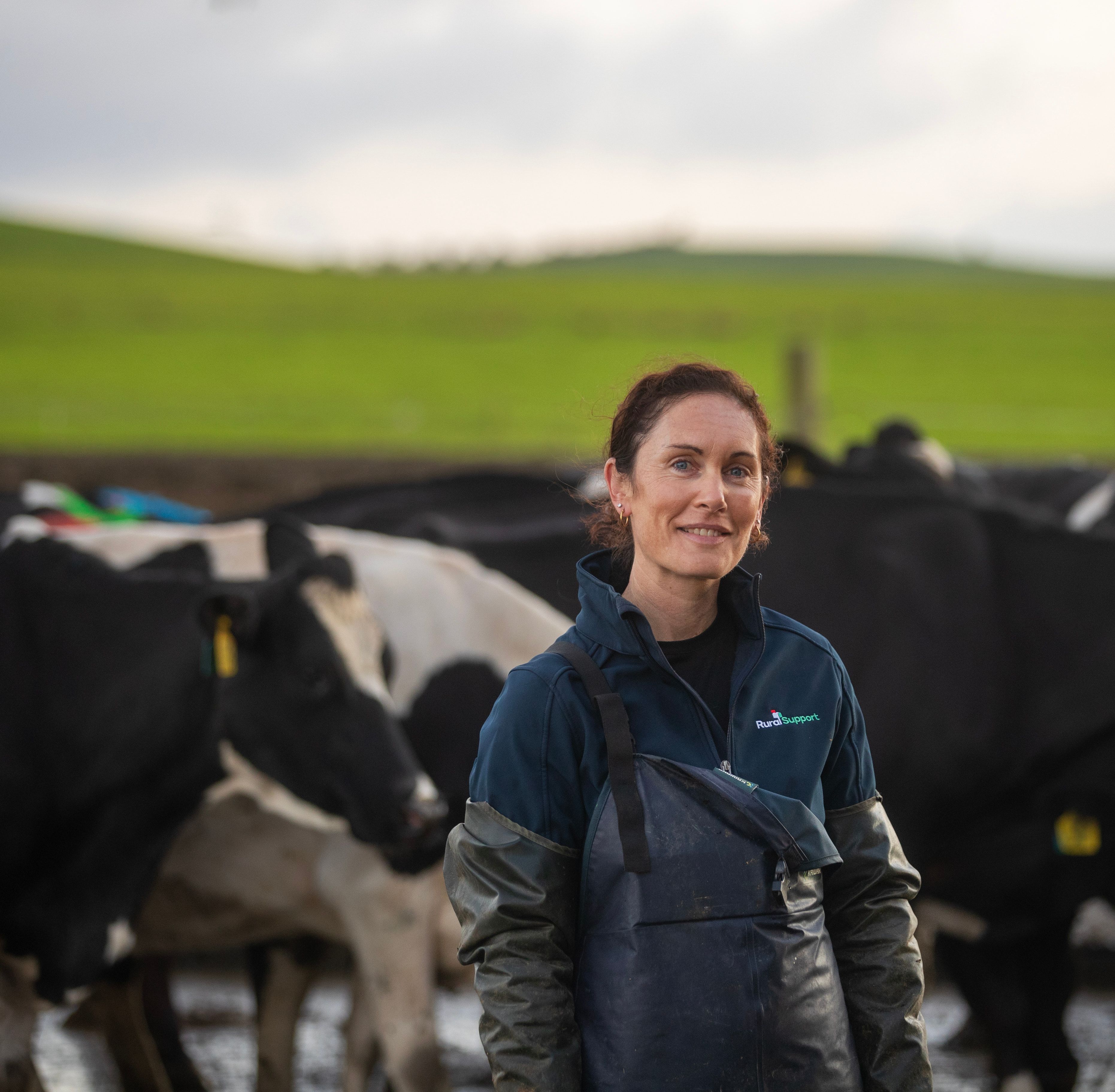 Woman near camera in farming gear with dairy cows behind. 