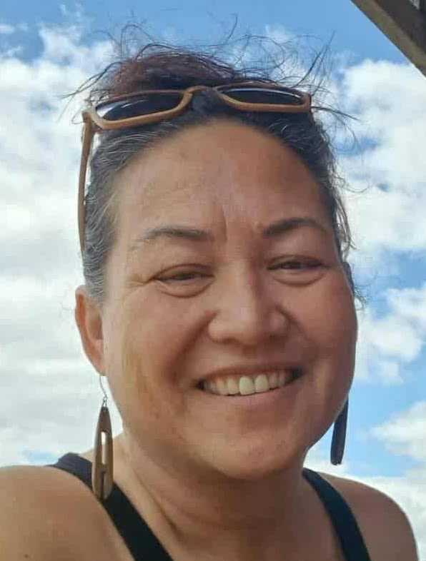 Head and shoulders image of smiling woman, with drop earrings and sunglasses on top of head. Blue sky with white clouds behind.