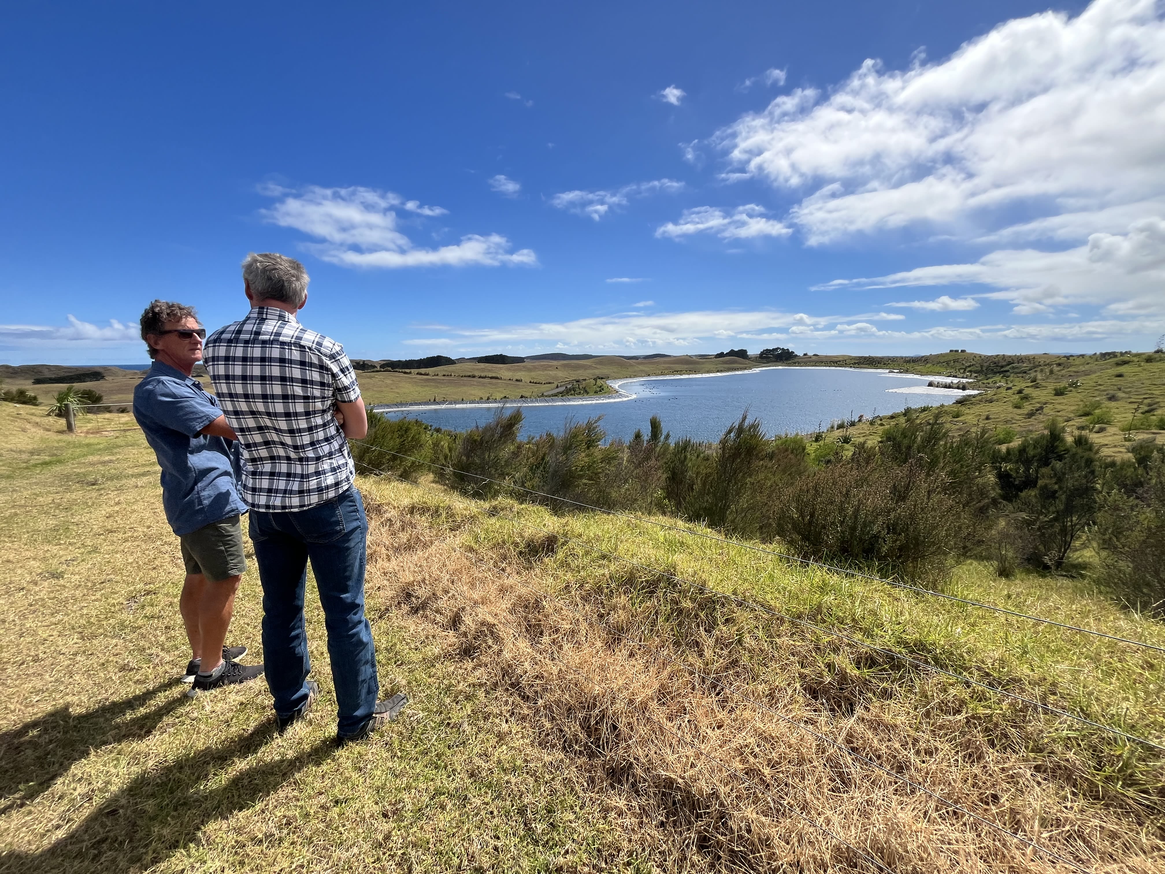 Two men stand talking on grassy hill, with bush and water reservoir in distance. 