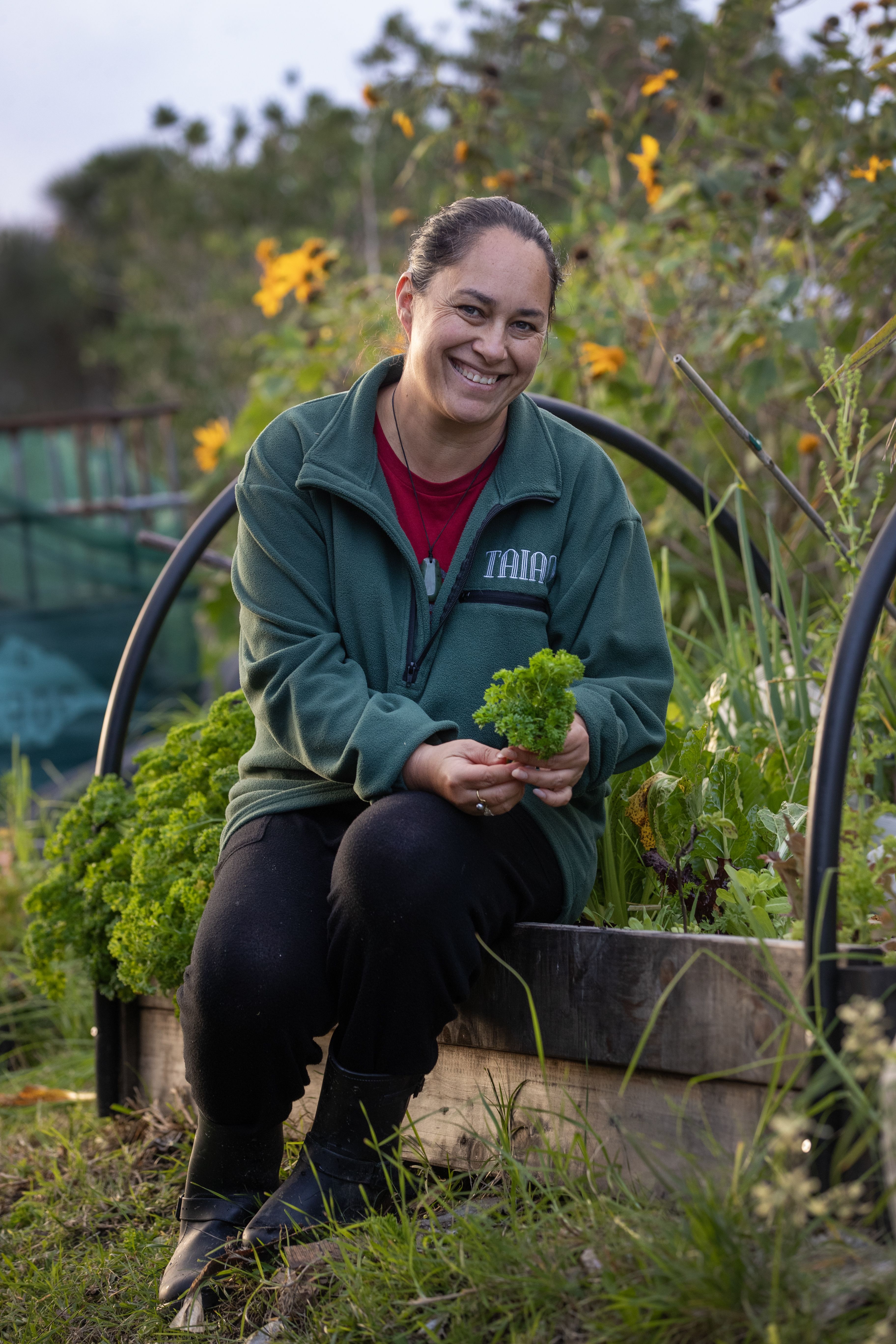 Smiling woman holding greenery in front of lush food gardens.