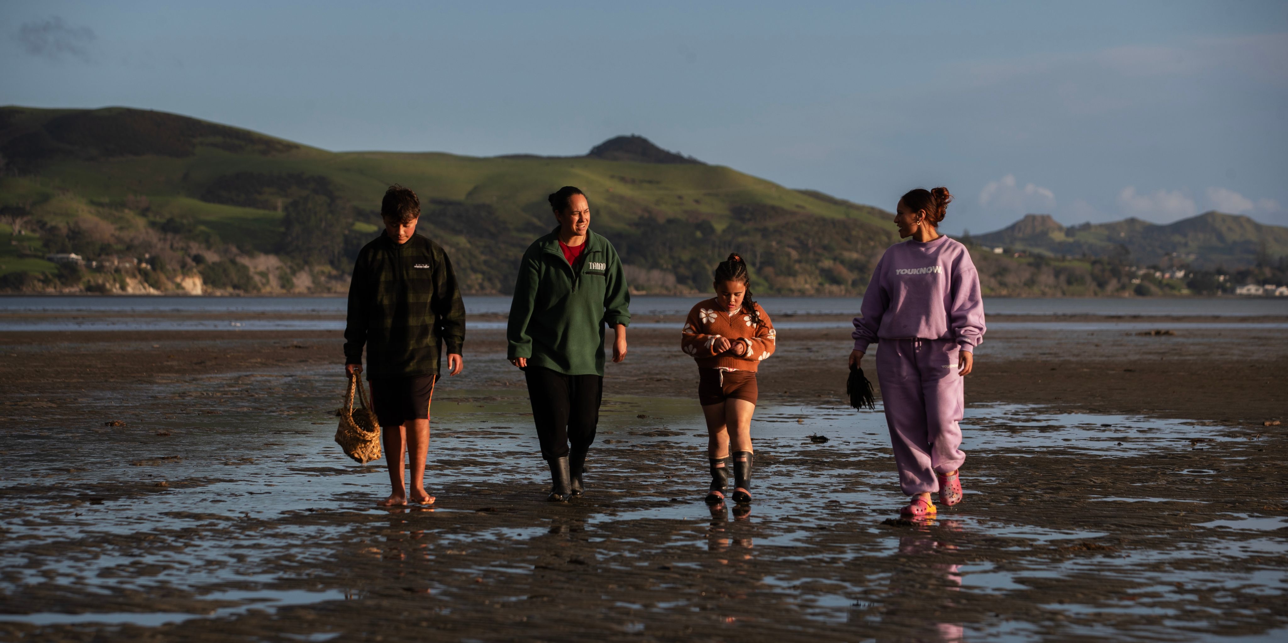 Four people walking across coastal mudflats with green hills behind, lad at left carrying a woven kete (basket)