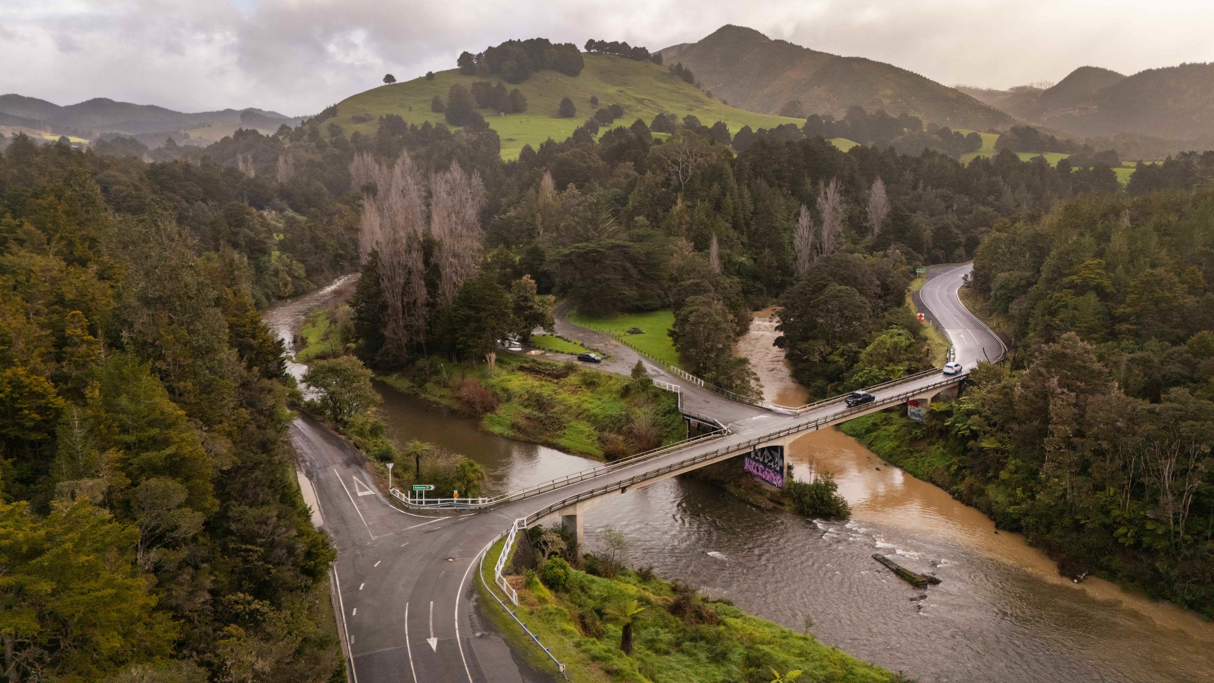 Aerial shot of road bridge crossing two rivers between thick forest with hills beyond. 