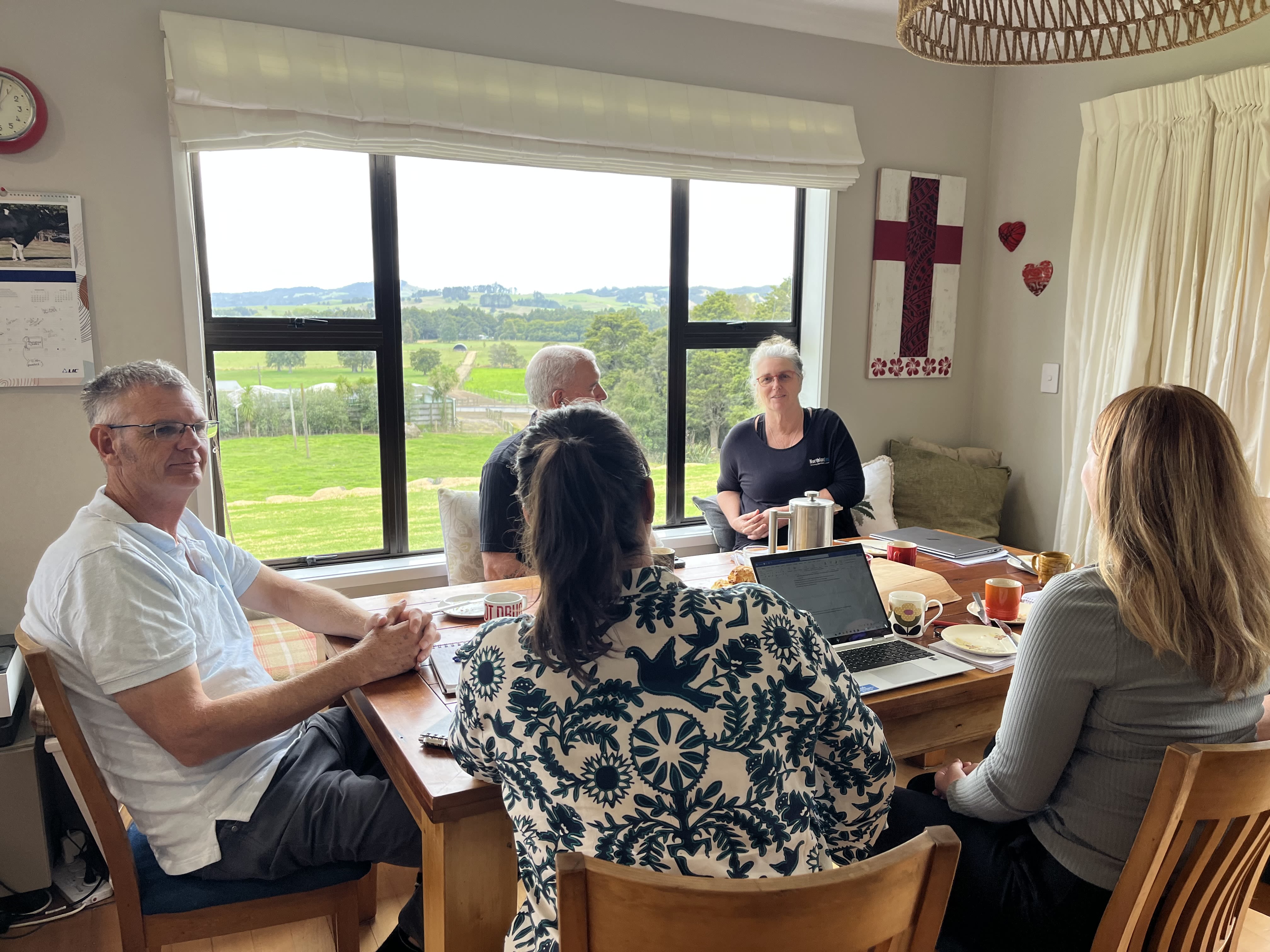 Five people sitting around a table, farm paddocks visible through the window,