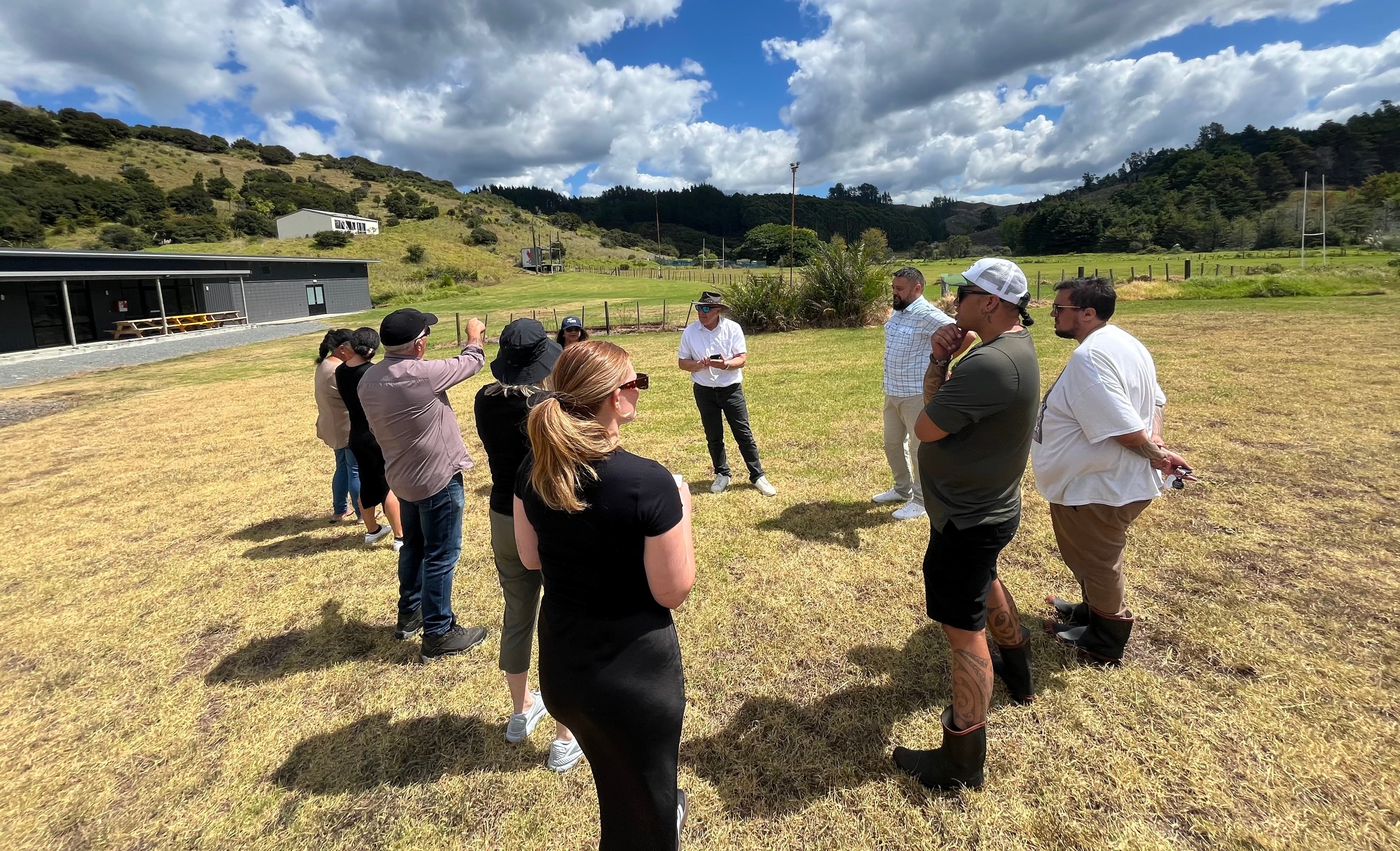 Group of people standing in open, listening to someone speaking. Sports facility to side.